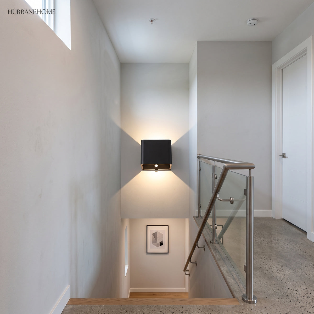 Black cube wall sconce lit on a stair wall beside a glass balustrade