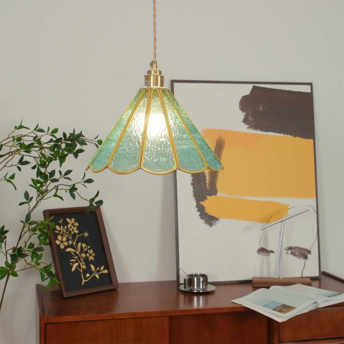 Blue glass pendant lit brightly above a desk with a framed botanical print