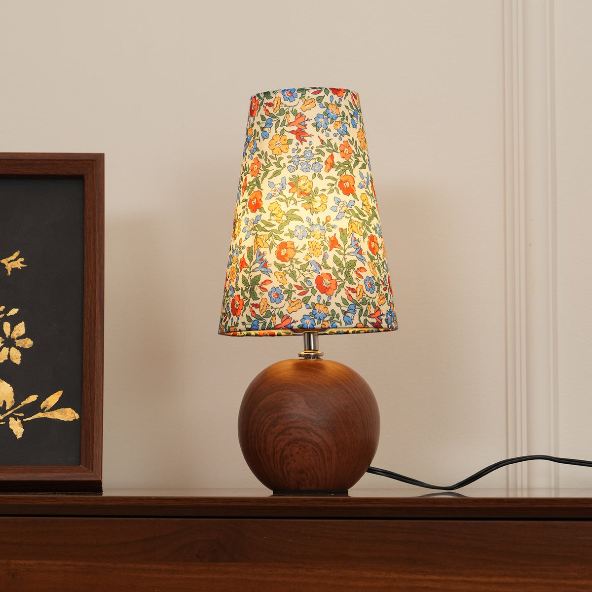 Warm floral shade table lamp lit beside a dark framed botanical picture