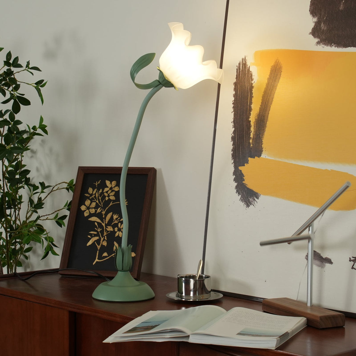 Flower table lamp lit on a teak sideboard beside a gold botanical framed print