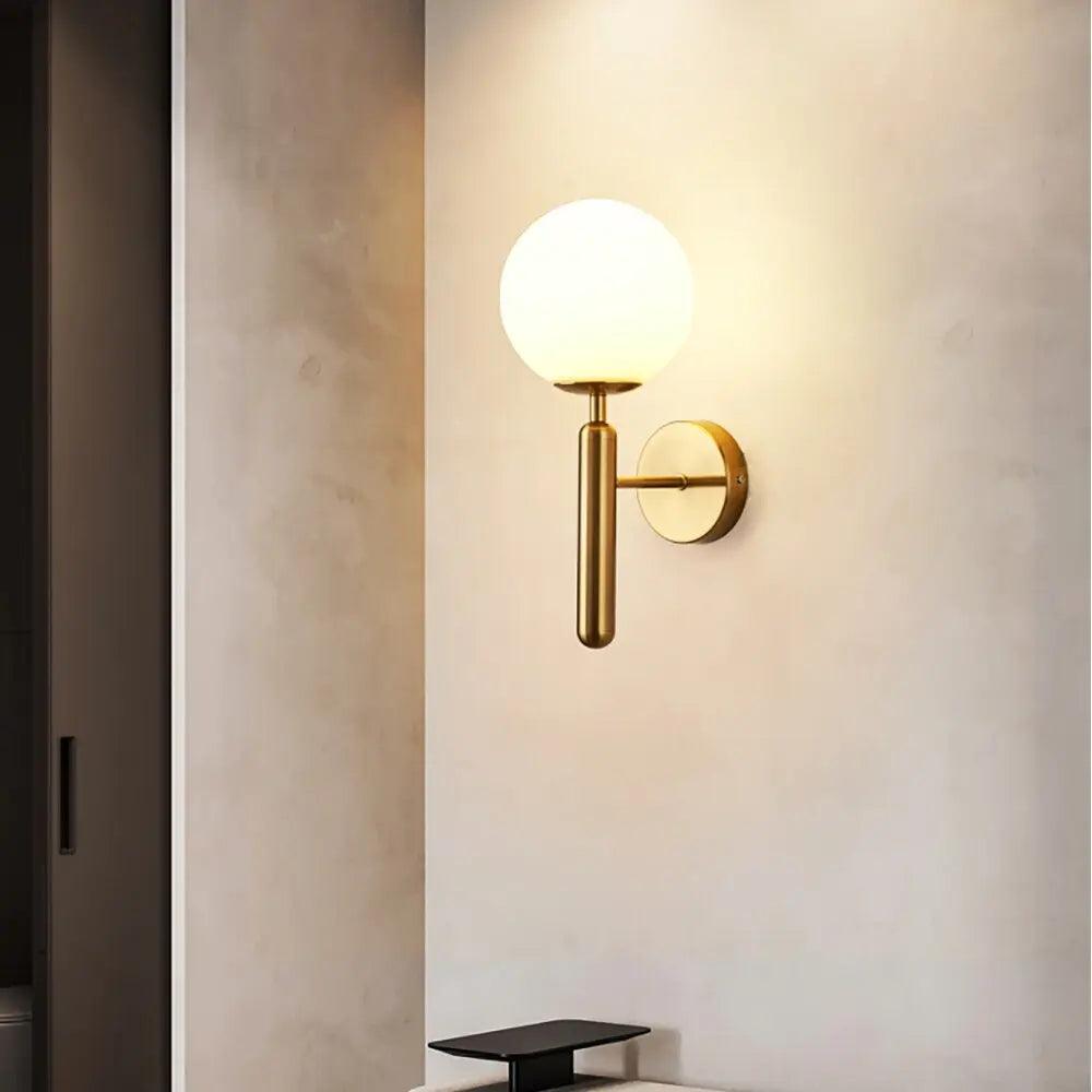 Gold sconce with a milk glass globe lit on a pale plaster wall
