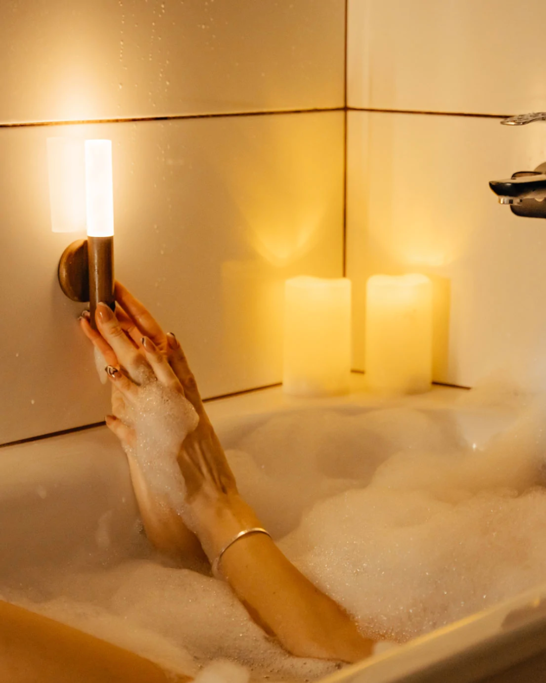 Hand reaching for the lit portable lamp beside a bath and candles