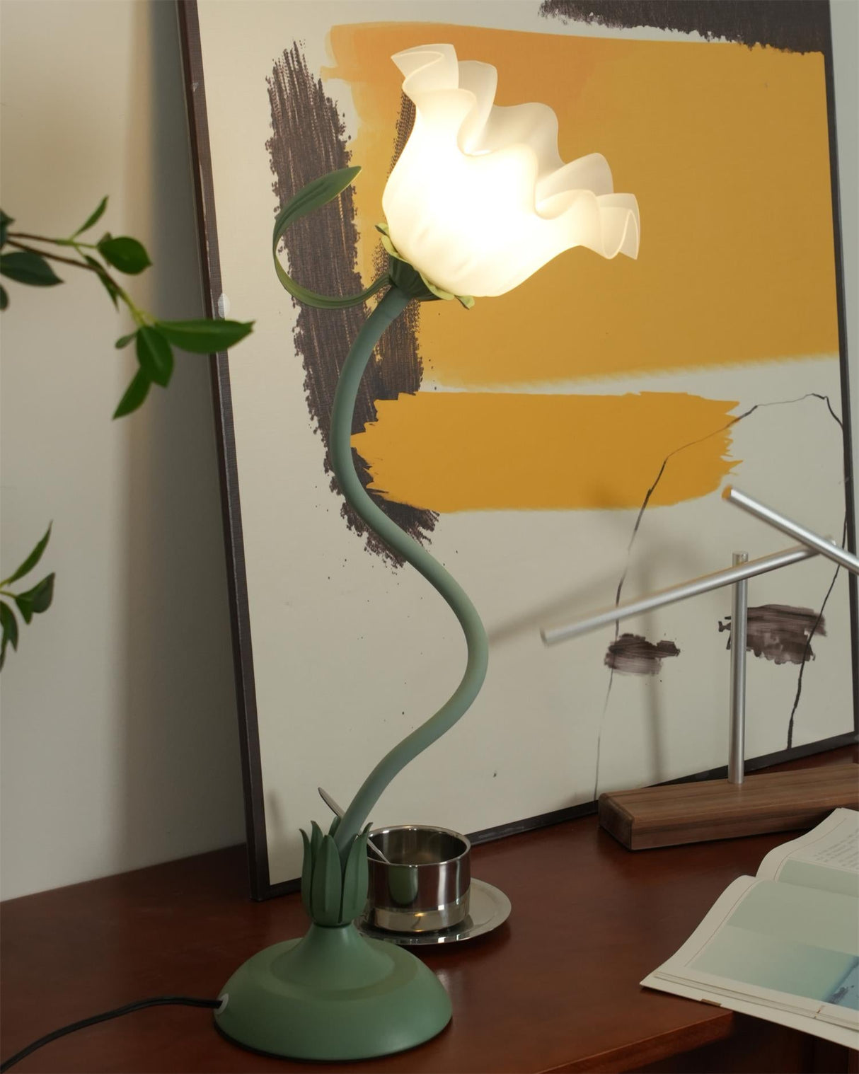 Tall flower table lamp lit on a sideboard with foliage and framed art behind