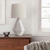 Diamond-shaped white ceramic lamp on a walnut sideboard beside a window