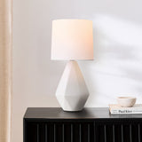 White ceramic geometric table lamp with linen shade on a dark sideboard