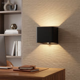 Black cube wall sconce lit on a textured wall above a desk