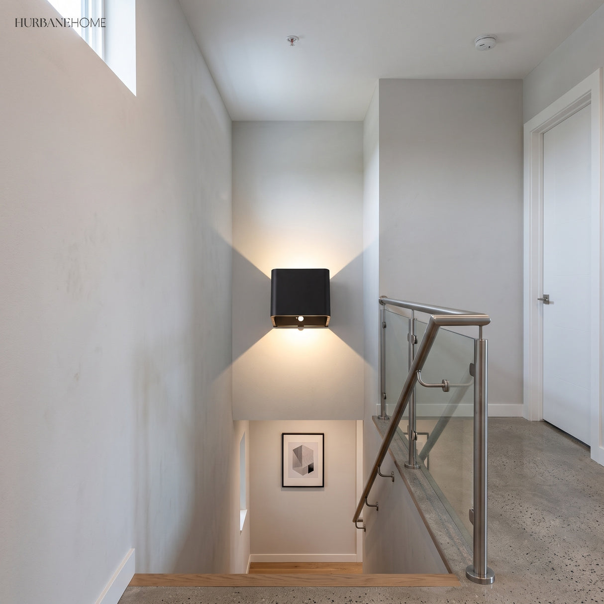 Black cube wall sconce lit on a stair wall beside a glass balustrade