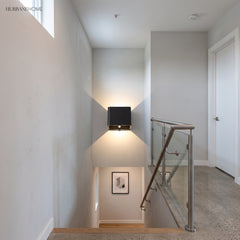 Black cube wall sconce lit on a stair wall beside a glass balustrade