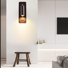 Black bedside reading light lit on a pale wall beside a wooden stool