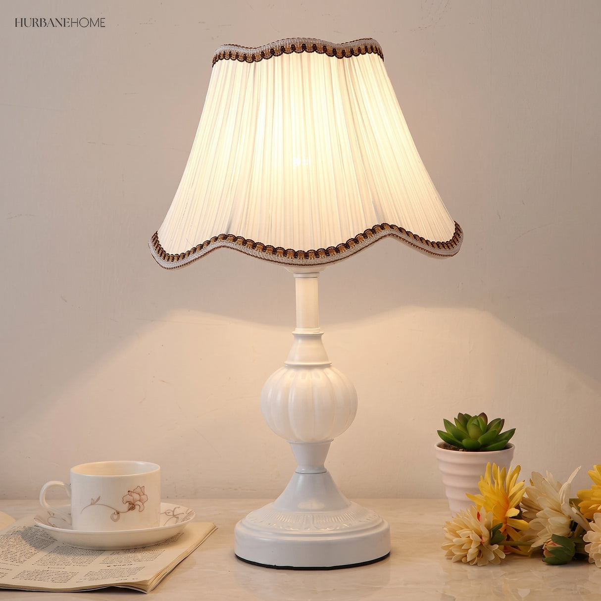 Lit bedside table lamp with white base beside a teacup and plant