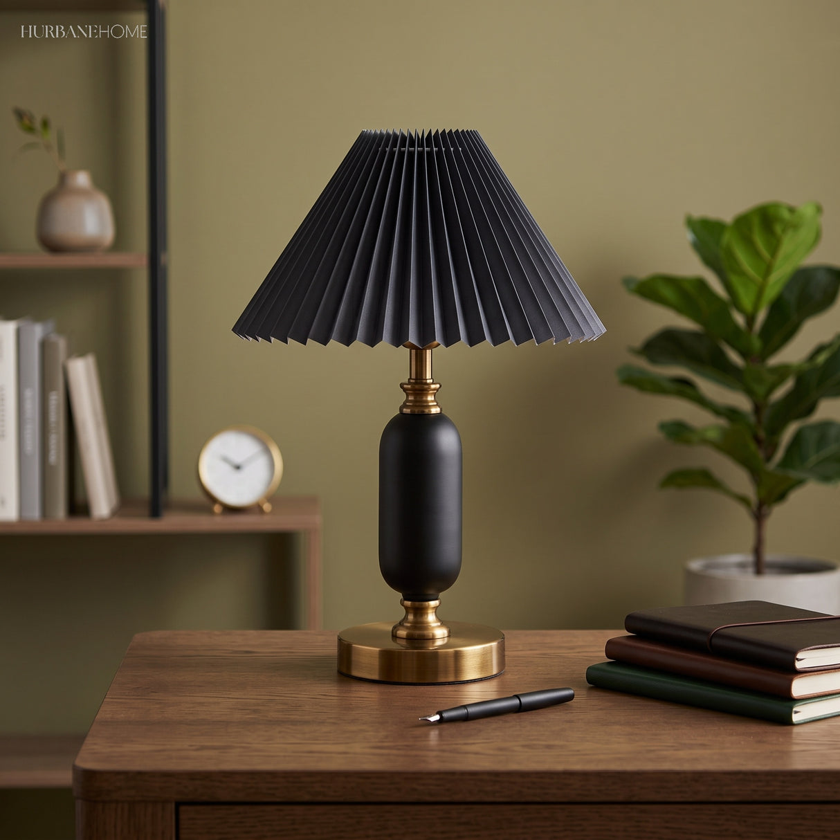 Black pleated table lamp with brass base on a desk against an olive wall
