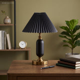 Black pleated table lamp with brass base on a desk against an olive wall