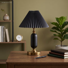 Black pleated table lamp with brass base on a desk against an olive wall