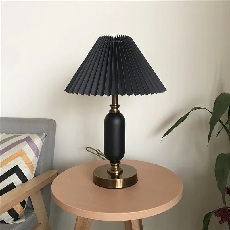 Black pleated table lamp with brass base beside a plant