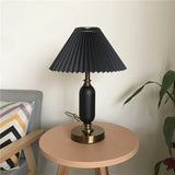 Black pleated table lamp with brass base beside a plant