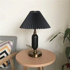 Black pleated table lamp with brass base beside a plant