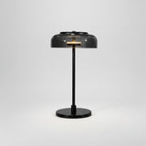 A modern table lamp with a black metal stand, round base, and a translucent dark gray dome shade, shown on a white background with its power cord visible.