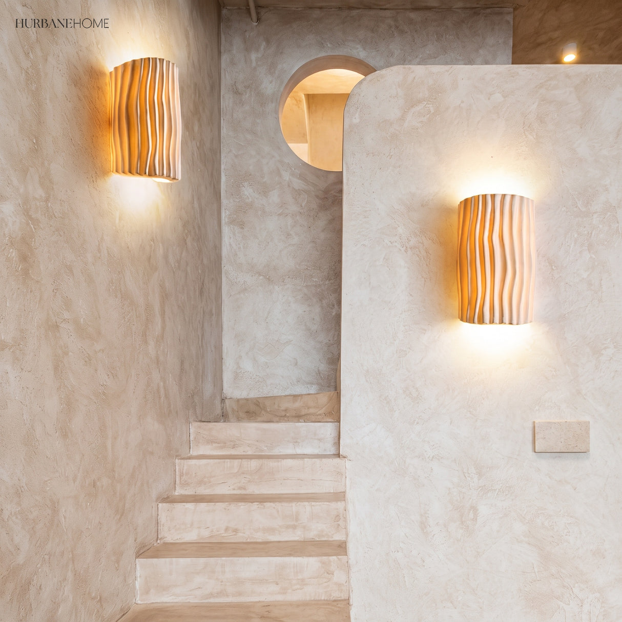 Two cream wall sconces lighting a plaster stairwell with a round window