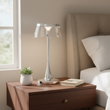 A modern table lamp with a clear glass shade and polished silver base, styled on a wooden nightstand next to a bed in a bedroom setting.