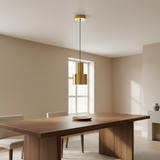 A modern dining room scene featuring a gold cylindrical pendant light hanging above a wooden dining table with neutral beige chairs.