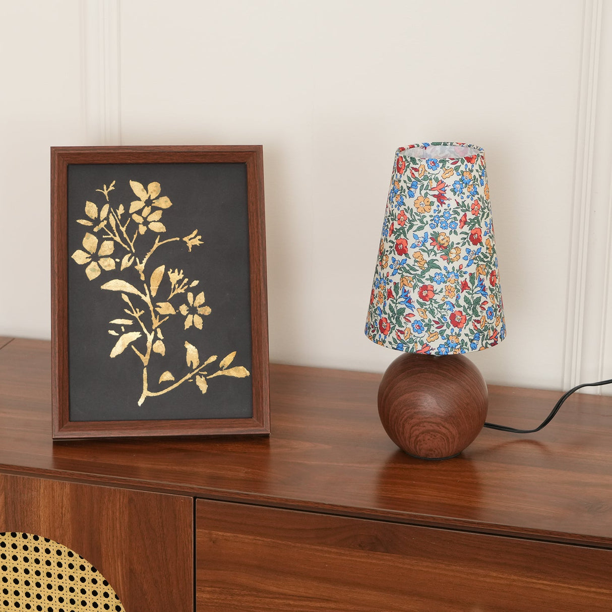 Floral lamp unlit on a walnut sideboard next to a framed leaf print