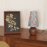Floral lamp unlit on a walnut sideboard next to a framed leaf print