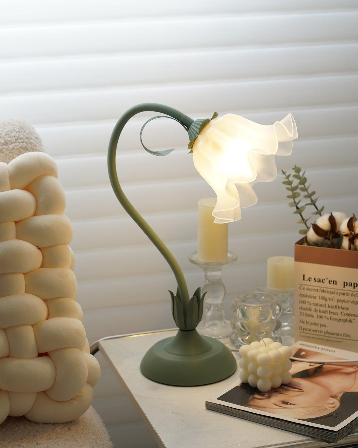 Flower table lamp lit above a candle holder and a stack of open magazines
