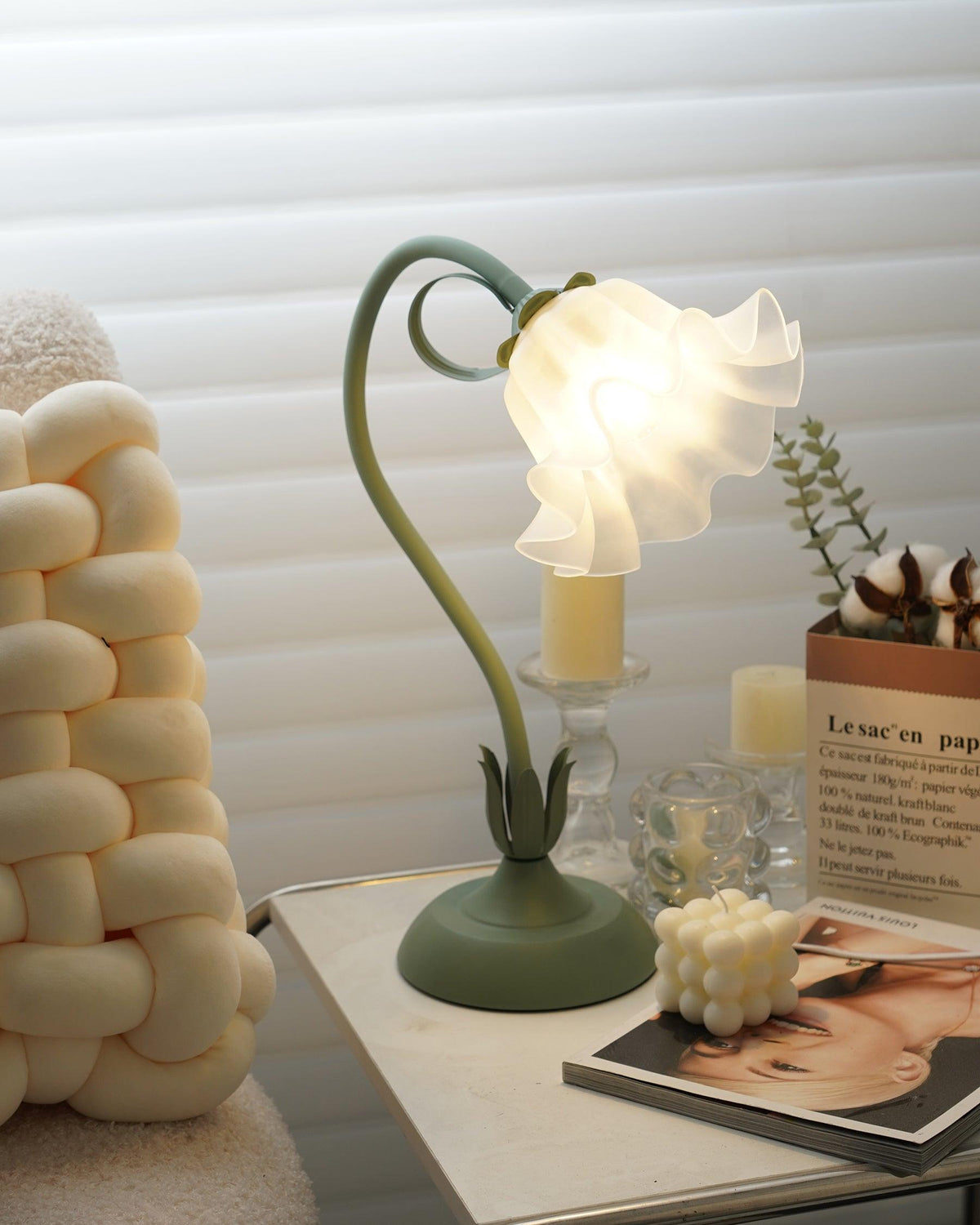Flower table lamp lit beside pillar candles and a knitted pouf on a side table
