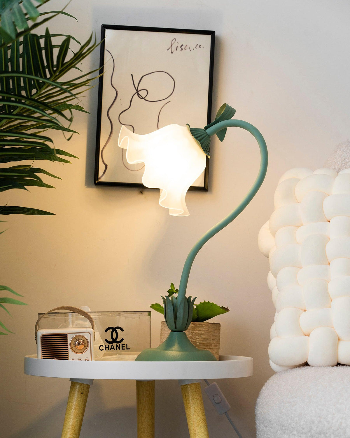 Flower table lamp casting warm light in a corner beside a palm and line art