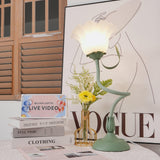 Upright flower table lamp lit beside dried stems and a stack of books