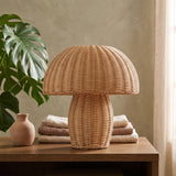 Natural rattan mushroom table lamp on a wood table beside a monstera leaf