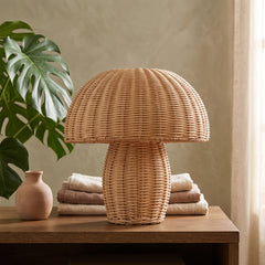 Natural rattan mushroom table lamp on a wood table beside a monstera leaf