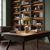 Gold dome lamp lit on a desk in front of a wall of lit timber shelving