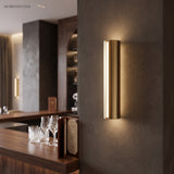 Gold tube sconce lit on a dark wall beside a timber cabinet