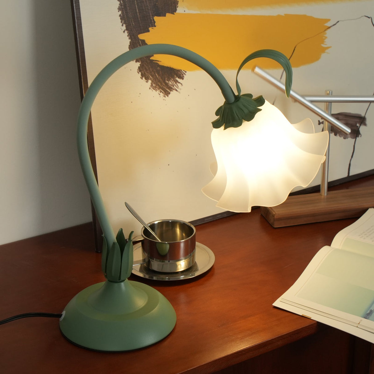 Green flower table lamp lit on a teak sideboard beside a brass cup and artwork