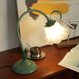 Green flower table lamp lit on a teak sideboard beside a brass cup and artwork