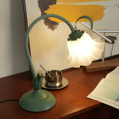 Green flower table lamp lit on a teak sideboard beside a brass cup and artwork