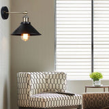 Black cone shade wall sconce lit on a green wall beside a window blind