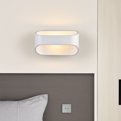 White oval sconce lit on a pale wall beside a bed