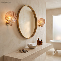 Two brass shell wall sconces flanking a round gold mirror in a warm bathroom