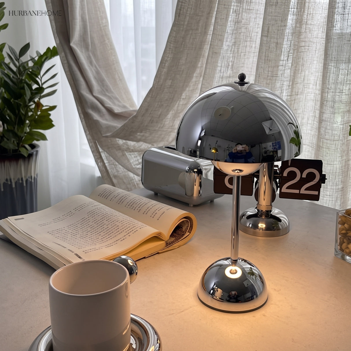 Chrome table lamp lit on a desk with an open book and mug by a window