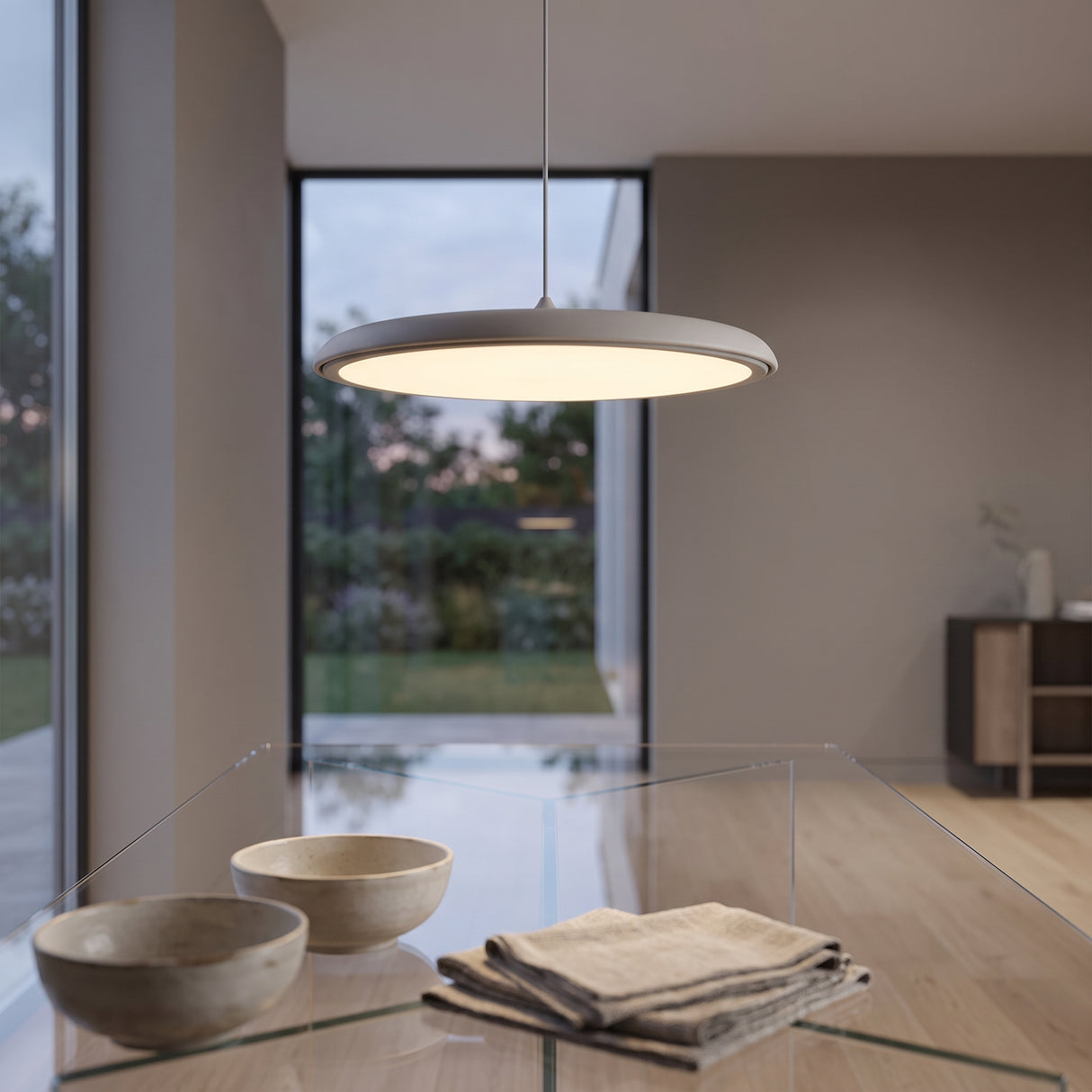 Grey disc pendant light glowing above a glass dining table with two ceramic bowls