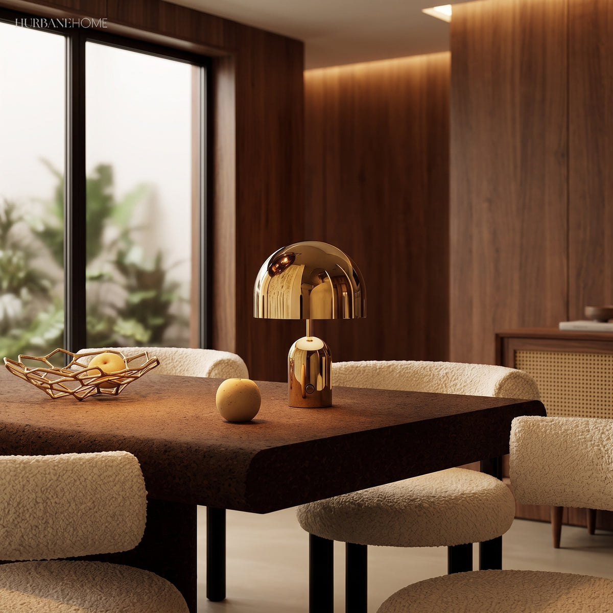 Gold table lamp lit on a dark dining table beside a fruit bowl and boucle chairs