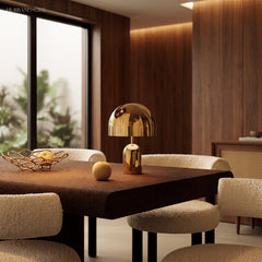 Gold table lamp lit on a dark dining table beside a fruit bowl and boucle chairs
