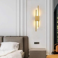 Gold linear sconce lit above a bed with a pale upholstered headboard