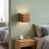 Wood cube wall sconce lit on a green wall beside a bed