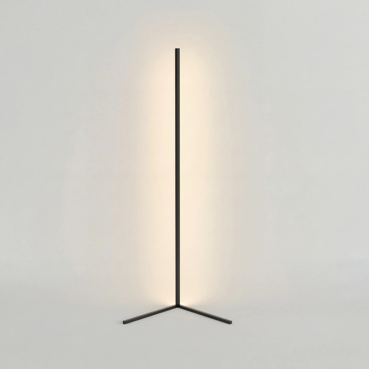 Black corner floor lamp on a triangle base with warm glow on a plain wall