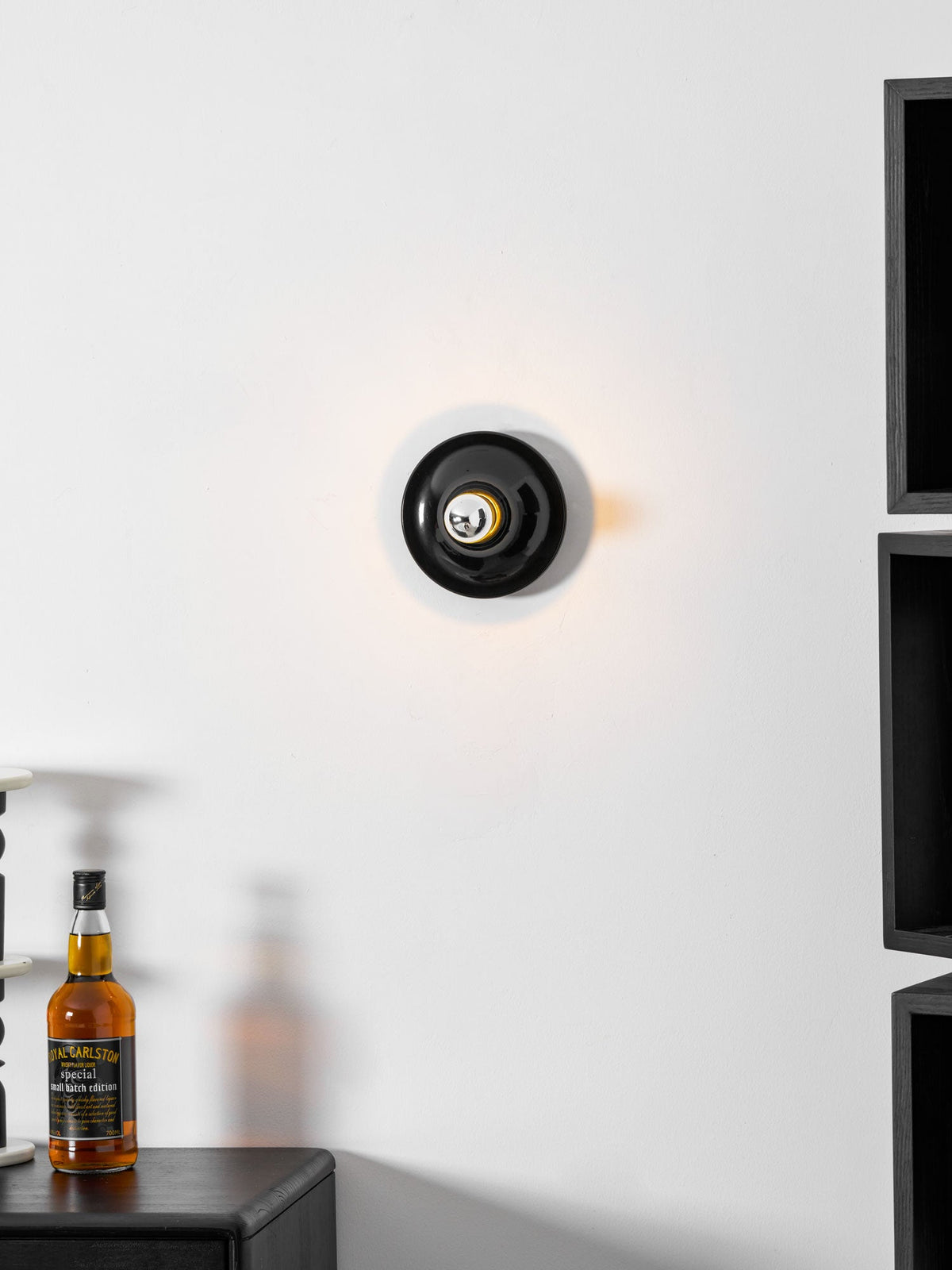 Black disc wall sconce seen face on above a dark cabinet and bottle