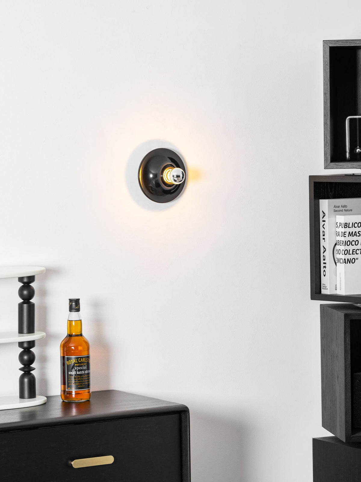 Black disc wall sconce lit above a dark cabinet holding glass bottles
