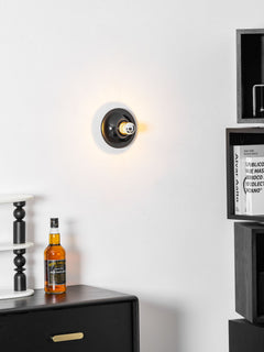 Black disc wall sconce lit above a dark cabinet holding glass bottles